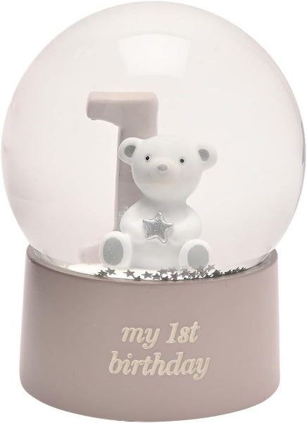 A first birthday snow globe figurine featuring a white teddy bear holding a silver star, sitting in front of a light gray number '1' on a beige base with 'my 1st birthday' written on it.