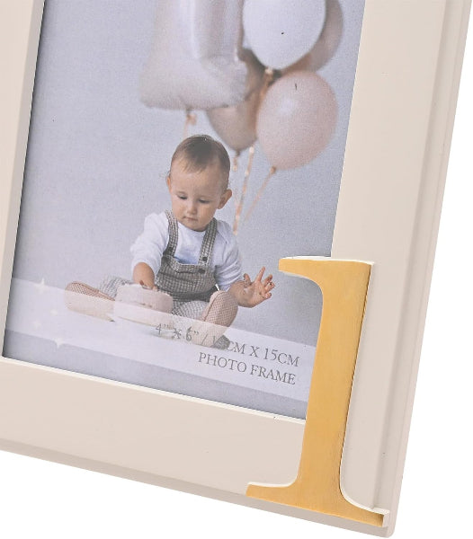 A cream-colored picture frame with a decorative gold letter 'J' on the lower right corner, holding a printed photo of a baby with balloons. The frame is shown at an angle against a white background.