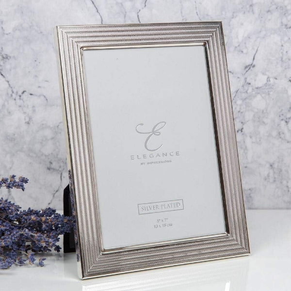 A silver-plated rectangular picture frame with a textured beaded border, standing on a white surface against a light gray marble-patterned background. The frame is shown empty, with a small decorative sprig of lavender on the left side.