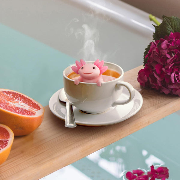 Pink axolotl-shaped tea infuser in a cup of tea on a wooden surface with a half-grapefruit and flowers.