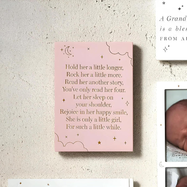 A pink square wall art print with a baby shower poem printed in black text, decorated with gold stars and clouds, displayed on a textured beige wall next to other baby photos and cards.