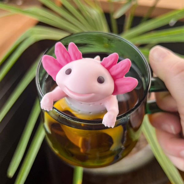 Pink axolotl-shaped tea infuser in a glass mug with tea, held by a hand with a plant in the background.