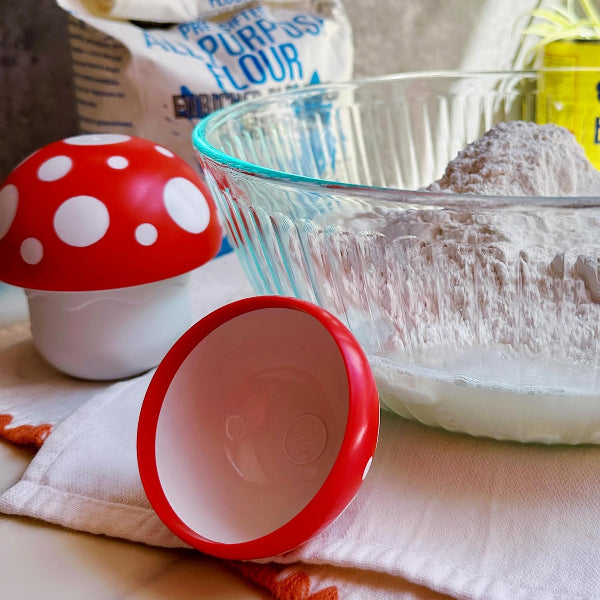 Red and white mushroom-shaped container with a clear glass bowl filled with flour on a kitchen counter.