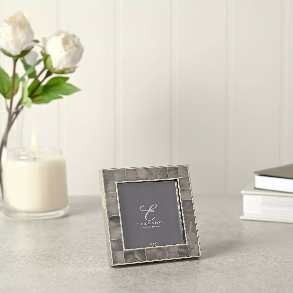 Silver photo frame with 'Elegance' branding on a light surface with a candle and flowers in the background.