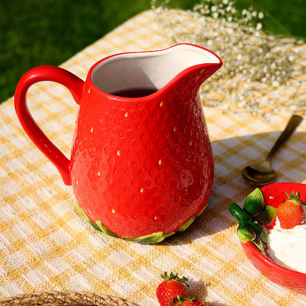 Red strawberry-shaped ceramic pitcher on a checkered tablecloth with a bowl of strawberries and cream.