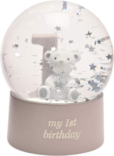 A clear glass or acrylic snow globe with a white teddy bear sitting in front of a large number 1, surrounded by silver star confetti. The base is a matte dusty pink with the words 'my 1st birthday' printed in a light pink script.