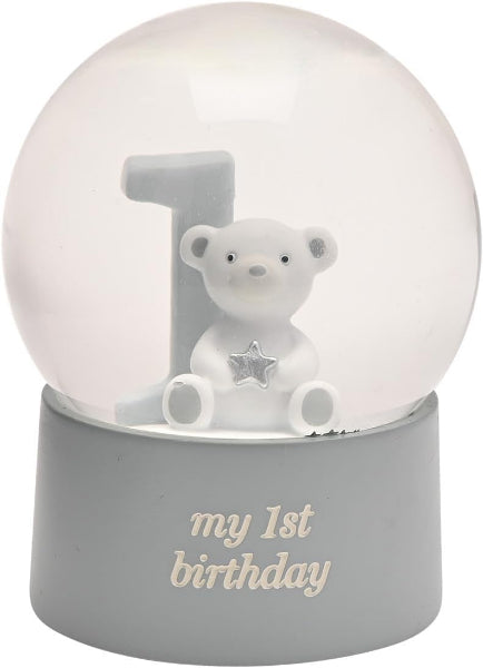 A first birthday snow globe figurine featuring a white teddy bear sitting in front of a gray number 1, with a silver star on its chest. The base is gray and printed with the words 'my 1st birthday' in a light beige font.