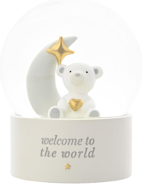 A white snow globe figurine featuring a teddy bear sitting on a crescent moon, both adorned with gold accents including a star on the moon and a heart on the bear's chest. The base is white with the text 'welcome to the world' in gray, and the entire piece is set against a solid white background.