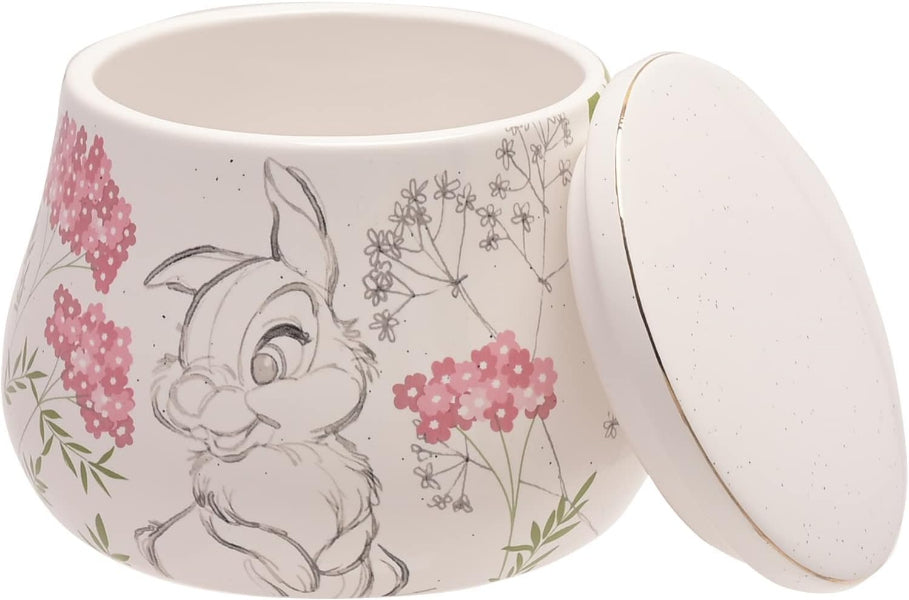 A white ceramic jar with its lid leaning against it, decorated with a sketch of a rabbit and pink flowers with green stems on a white background.