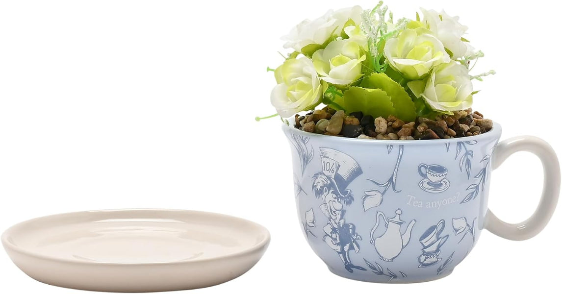 A light blue ceramic teacup planter with a white handle, decorated with blue Alice in Wonderland illustrations, filled with artificial white roses and green leaves. A separate matching light beige saucer is shown to the left on a plain white background.