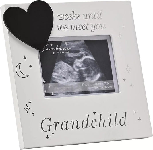 A white square baby photo frame with a black heart-shaped ornament attached to the top left corner. The frame features printed text reading 'weeks until we meet you' at the top and 'Grandchild' at the bottom, along with small star and moon illustrations.