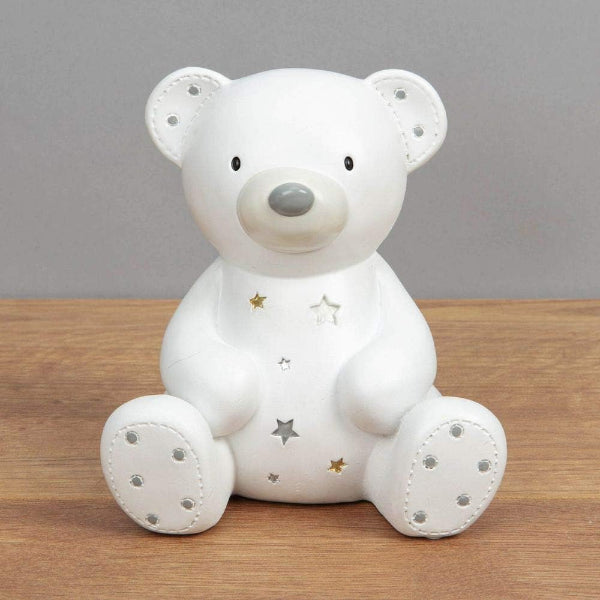 A white ceramic teddy bear figurine sitting on a wooden surface against a gray background, featuring star cutouts on its chest and soles of its feet.