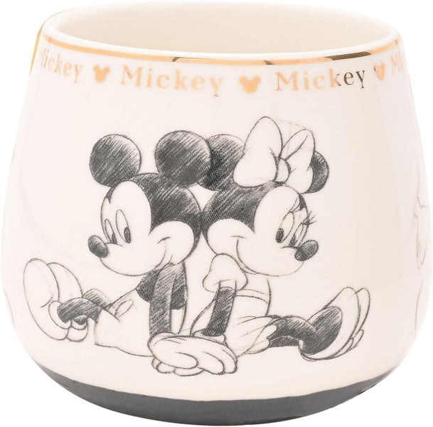 An off-white ceramic cup featuring a black sketch-style illustration of Mickey Mouse and Minnie Mouse sitting together, with 'Mickey' text and heart icons printed around the rim.