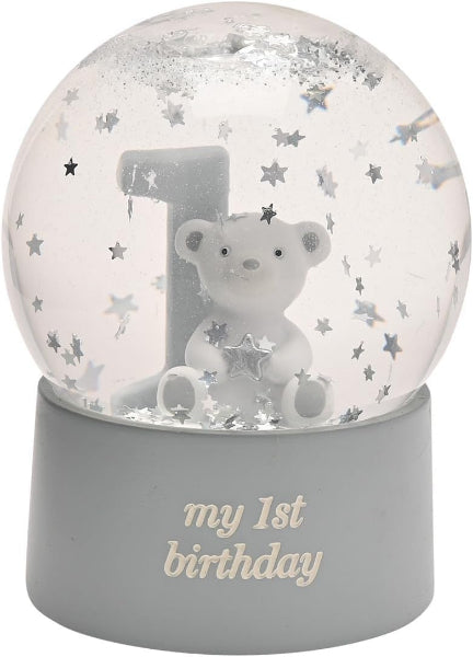 A first birthday snow globe featuring a white teddy bear, a large number 1, and silver star confetti inside a clear globe, set on a gray base with 'my 1st birthday' written in gold lettering.