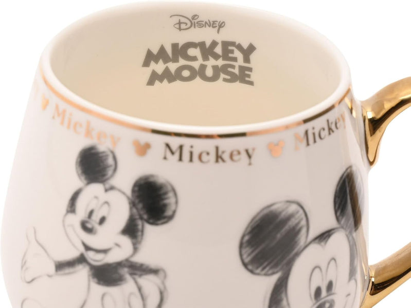 A close-up of a Disney Mickey Mouse ceramic mug with a white body, black and gray Mickey illustrations, gold text and trim, and a gold handle on a white background.