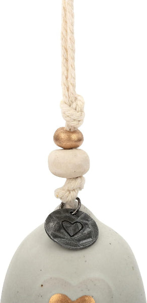 A close-up of a hanging ornament or decorative bead strand with a cream rope, a small metallic gold bead, a round off-white bead, a dark metal tag engraved with a heart, and the top of a pale gray ceramic or stone object visible below.