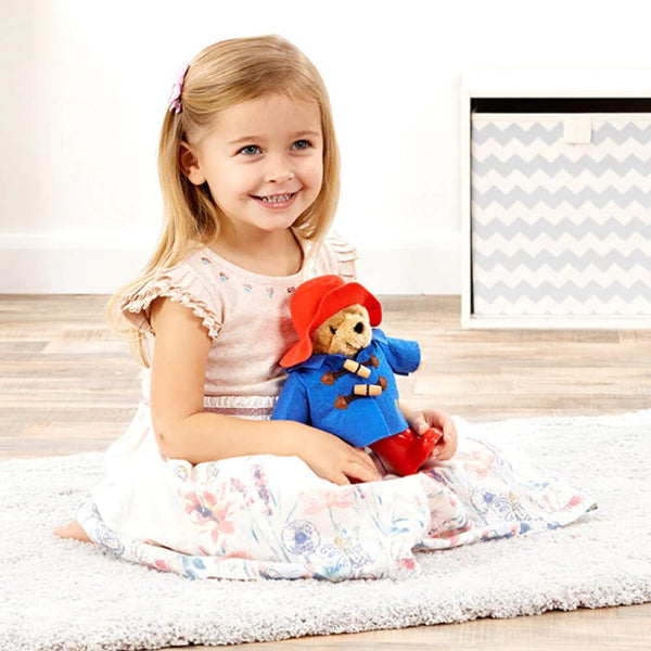 Young girl holding a teddy bear with a red hat and blue coat on a white rug.