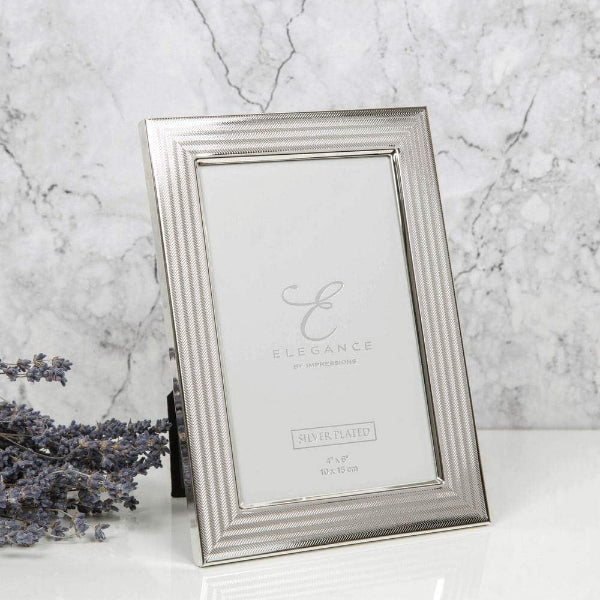 A silver picture frame with a textured, ribbed border and a clear glass insert, standing on a white surface against a gray marble background. A sprig of dried lavender is visible on the left side.