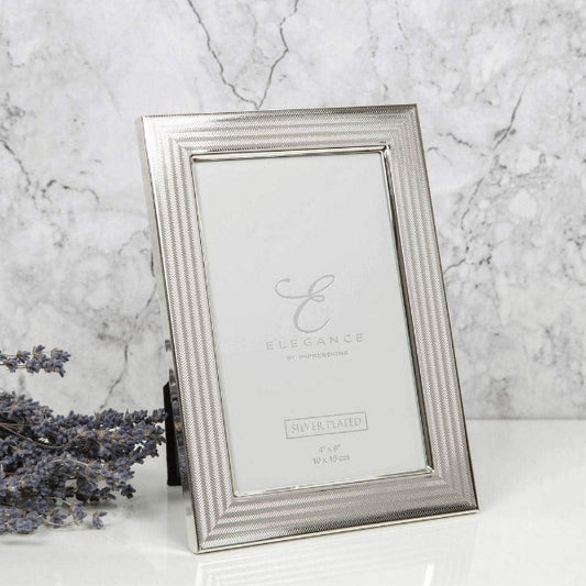 A silver picture frame with a textured, ribbed border and a clear glass insert, standing on a white surface against a gray marble background. A sprig of dried lavender is visible on the left side.