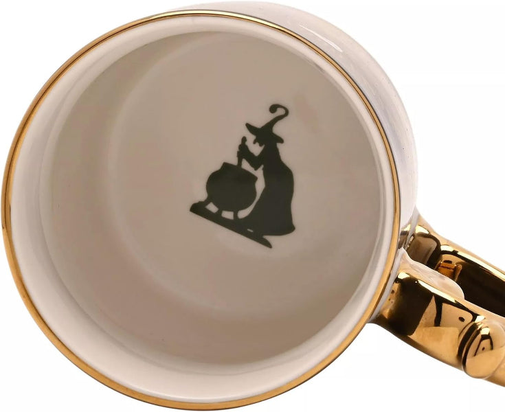 Close-up of a white ceramic mug with a gold rim and gold handle, featuring a black silhouette of a witch baking a cauldron inside the mug.