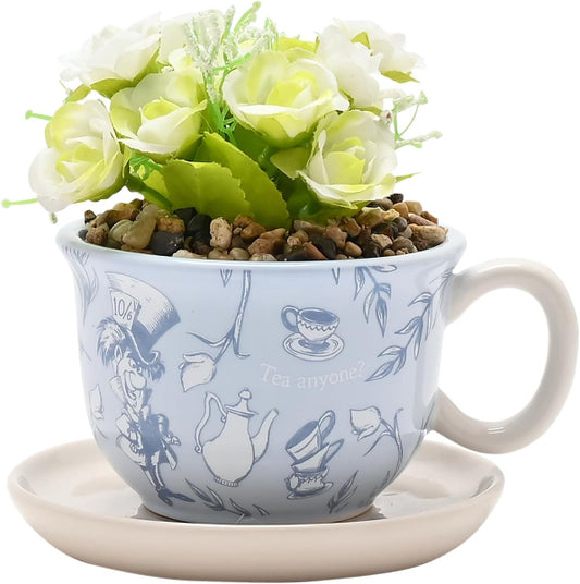 A decorative ceramic teacup planter with a matching saucer, filled with faux white roses and green leaves over small pebbles. The cup is light blue with blue illustrations of teapots, cups, and floral motifs, plus the words 'Tea anytime' and '100%'.
