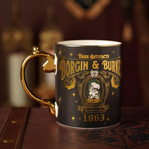 A dark ceramic mug with a gold handle and gold printed gothic-style artwork reading 'Borgin & Burlesque' and 'Dark Artifacts,' set on a brown leather surface with a blurred warm background.