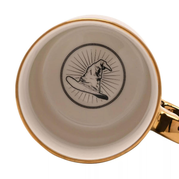 Close-up of the inside of a ceramic mug with a gold rim, a gold handle, and a black printed illustration of a pointed hat inside a circular frame on a white background.