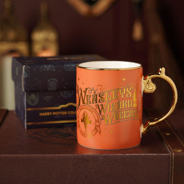 An orange ceramic Harry Potter mug with gold foil lettering reading 'Wizards of the Coast' and a gold handle, shown in front of its dark blue branded box on a brown surface