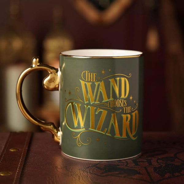 A dark green ceramic mug with a gold metallic handle and the phrase 'The Wand Chooses the Wizard' printed in gold lettering on the front, set on a dark wooden surface with a blurred background.