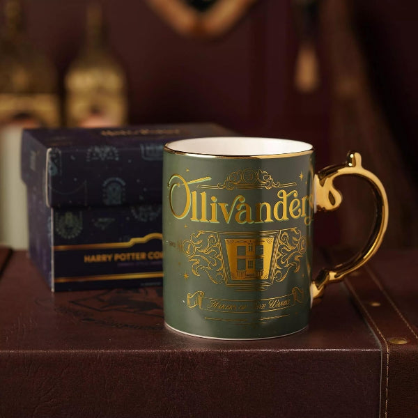 A dark green ceramic mug with gold foil detailing reading 'Ollivander' and the Hogwarts crest, shown with its branded blue gift box on a brown leather surface.
