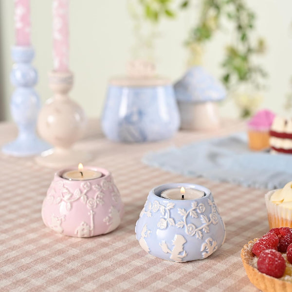Two lit candles in decorative holders on a table with pastel decor and food items.