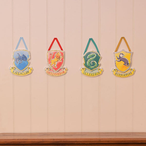 Four house crests from Harry Potter on a wooden shelf against a beige wall.