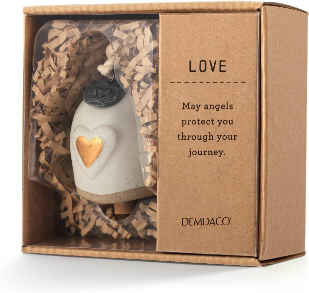 A ceramic angel ornament by Demdaco inside its original brown cardboard gift box with crinkle paper filler. The ornament is off-white with a gold heart and a dark grey rose detail.
