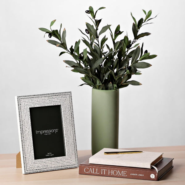 Decorative photo frame, vase with greenery, and book on a wooden surface.