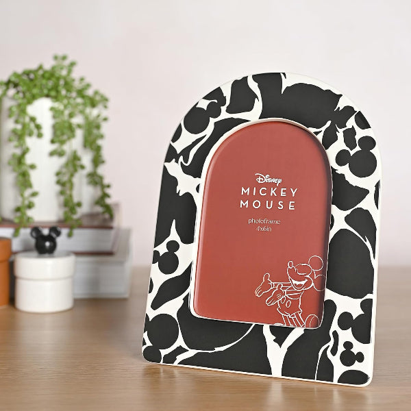 A Mickey Mouse themed photo frame with a black and white cow print pattern, standing on a wooden surface with a blurred background of a plant and books.