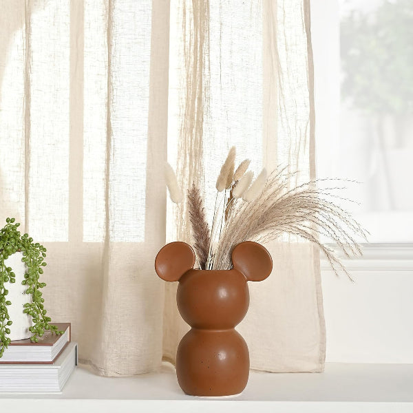 Brown vase with mouse ears on a surface with a curtain and plant in the background