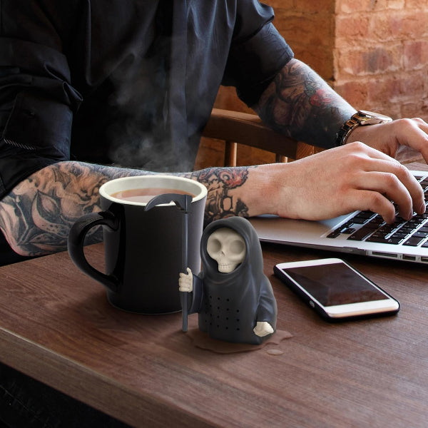 Person using a laptop with a steaming mug and skeleton-shaped cup holder on a wooden desk.