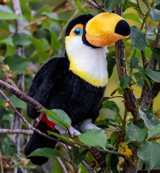 A plush toy toucan perched on a tree branch among green leaves, with a large yellow and orange beak, black body, white chest, blue eye, and small red accent near the tail.