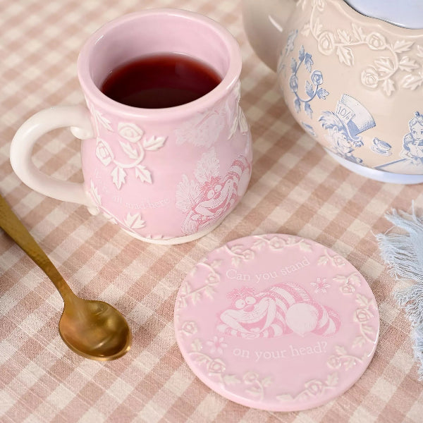 Pink mug with floral design, matching coaster, and teapot on a checkered tablecloth.