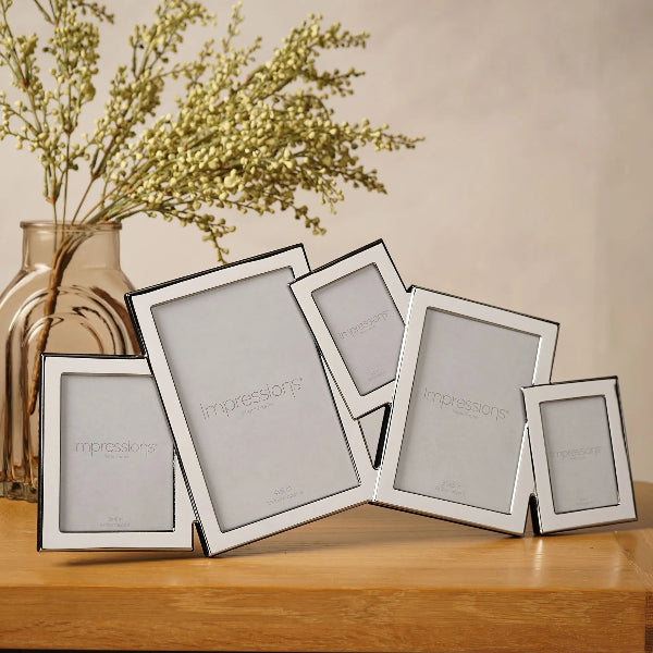 A set of six silver picture frames in different sizes, arranged on a wooden table next to a glass vase with dried flowers. The frames have a polished silver finish and display the brand name 'Impressions' on the inner card.