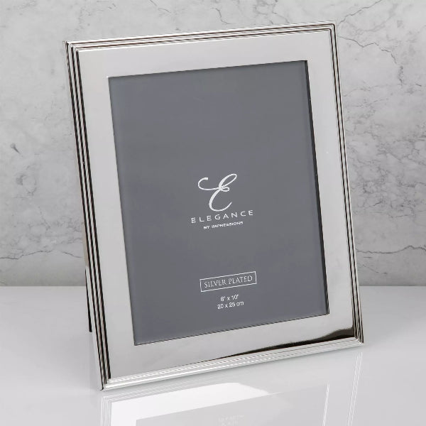 A silver-plated picture frame with a standing easel design, shown on a white reflective surface against a light gray marble background. The frame is empty, displaying a dark gray placeholder insert with the brand name 'ELEGANCE' and size details.