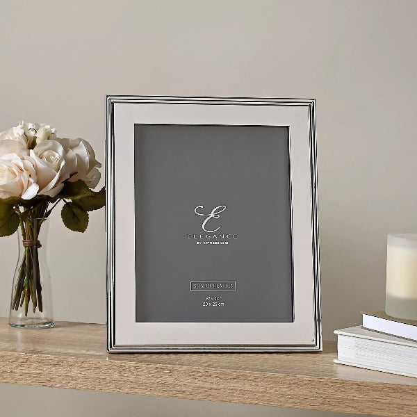 A silver picture frame with a white mat and thin dark edge lines, standing on a wooden shelf. The frame is branded with 'Elegance by Impression' and is styled with a vase of white roses on the left and a stack of books with a candle on the right against a neutral beige wall.
