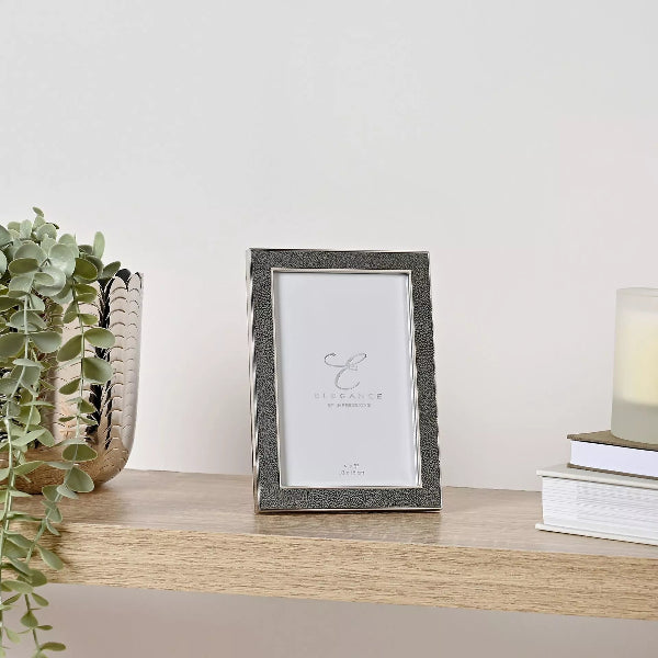 A vertical rectangular picture frame with a textured dark gray frame and a white insert, displayed on a light wood shelf next to a potted eucalyptus plant and a stack of books with a candle.