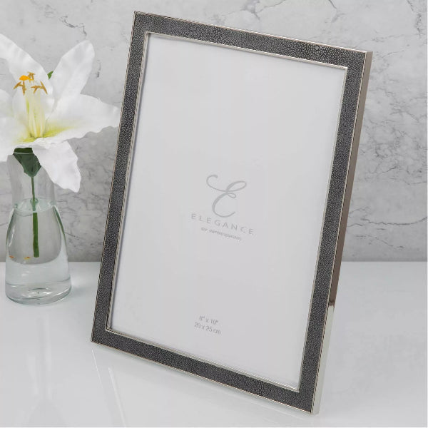 A rectangular picture frame with a textured silver border and a white insert, shown standing on a white surface beside a small glass vase with a white flower. The center of the insert reads Elegance.