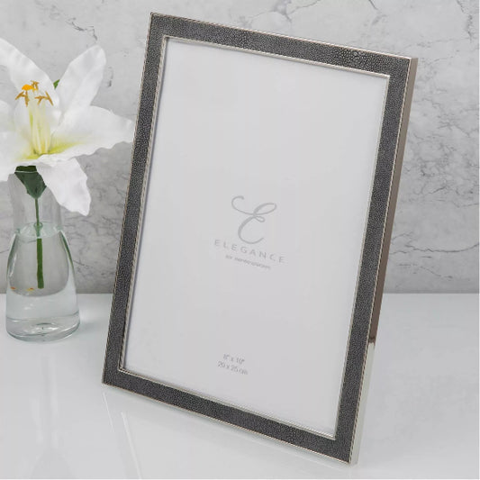 A rectangular picture frame with a textured silver border and a white insert, shown standing on a white surface beside a small glass vase with a white flower. The center of the insert reads Elegance.